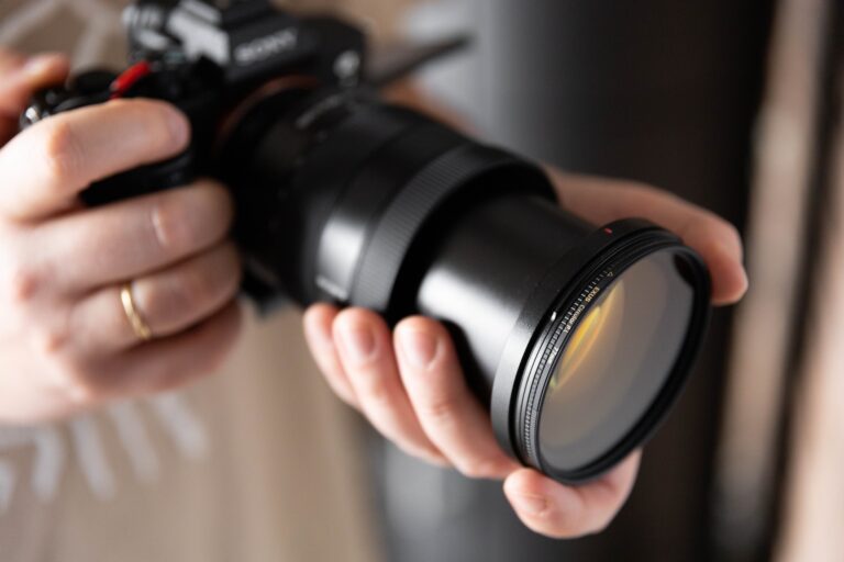 Dłonie trzymają czarny aparat fotograficzny z dużym obiektywem. Na obiektyw założony jest okrągły filtr fotograficzny z żółtawym odcieniem.