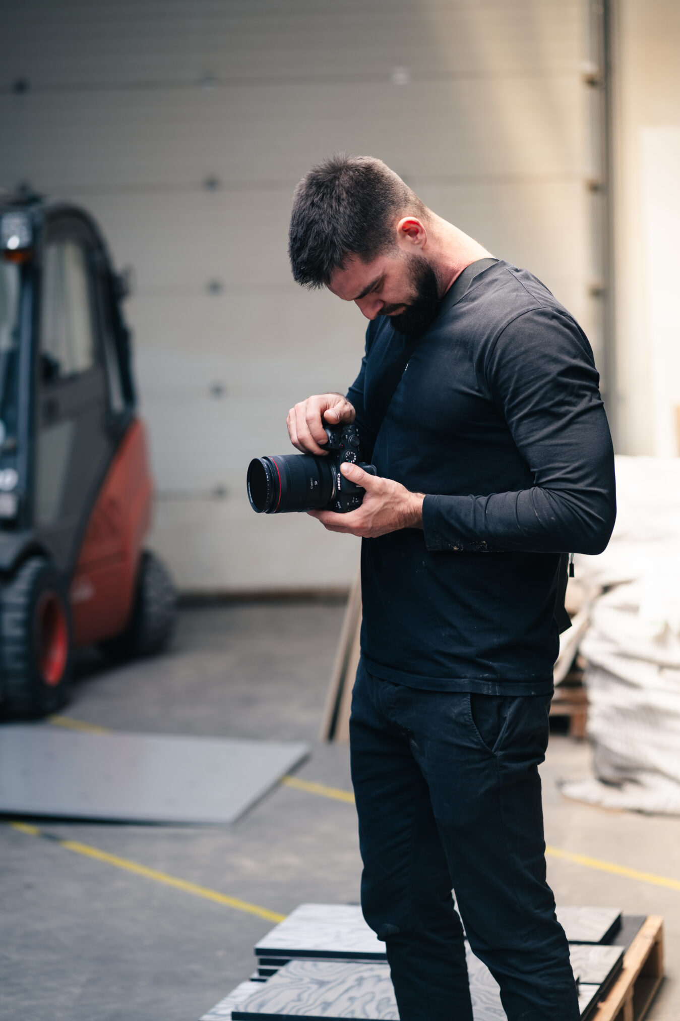 Mężczyzna w czarnej koszulce z długim rękawem trzyma aparat fotograficzny i patrzy na jego wyświetlacz. Stoi w pomieszczeniu przypominającym warsztat lub garaż z betonową podłogą i samochodem w tle.