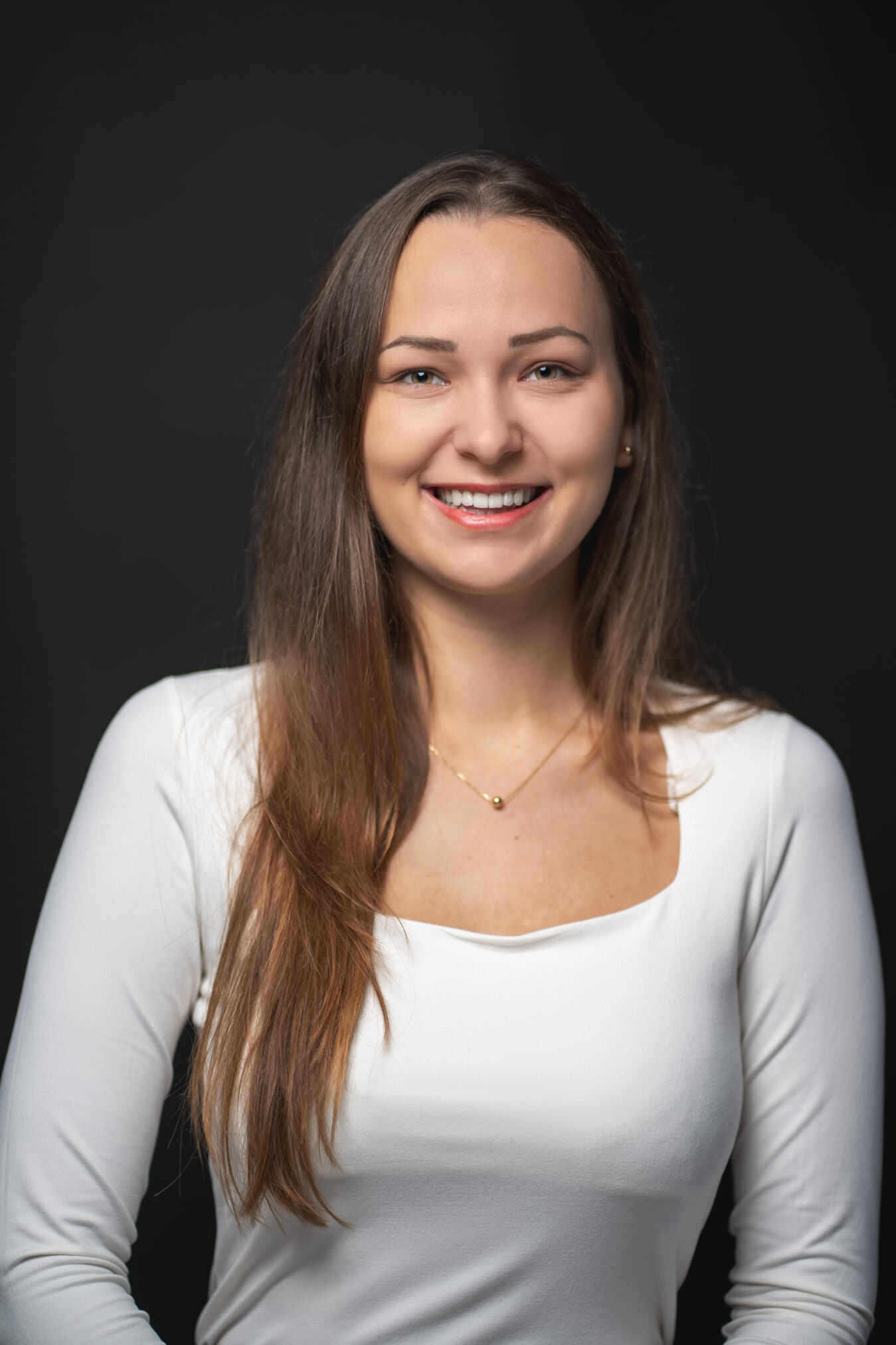 Młoda kobieta z długimi brązowymi włosami uśmiecha się szeroko do kamery, ubrana w białą bluzę z długimi rękawami. Zdjęcie wykonane na ciemnym tle w stylu portretowym.