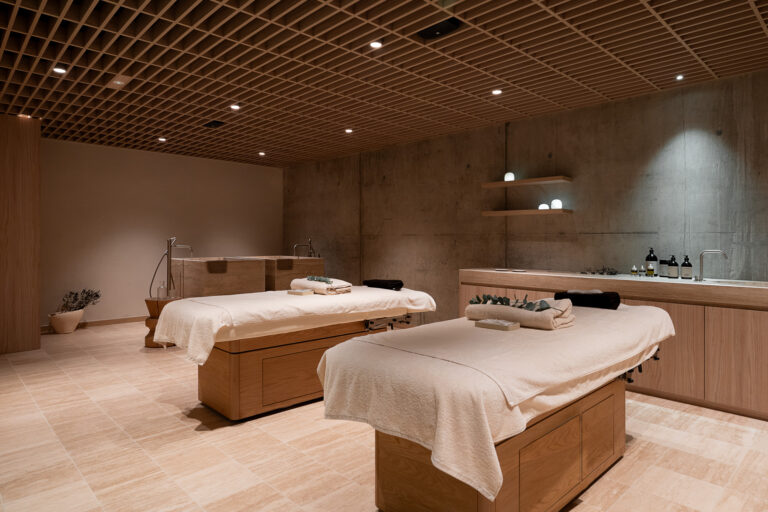 Luksusowe spa z dwoma drewnianymi łóżkami do masażu przykrytymi białymi ręcznikami. Wnętrze utrzymane w ciepłych, naturalnych kolorach z drewnianym sufitem kratowym, betonowymi ścianami i wbudowanymi półkami z kosmetykami.