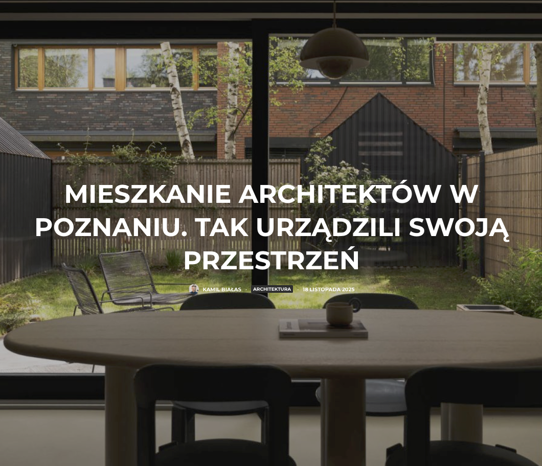 Widok z wnętrza mieszkania przez duże okna na nowoczesne budynki mieszkalne z cegły i drewna, otoczone zielenią i brzozami. Na pierwszym planie drewniany stół z czarnymi krzesłami i wiszącą lampą.