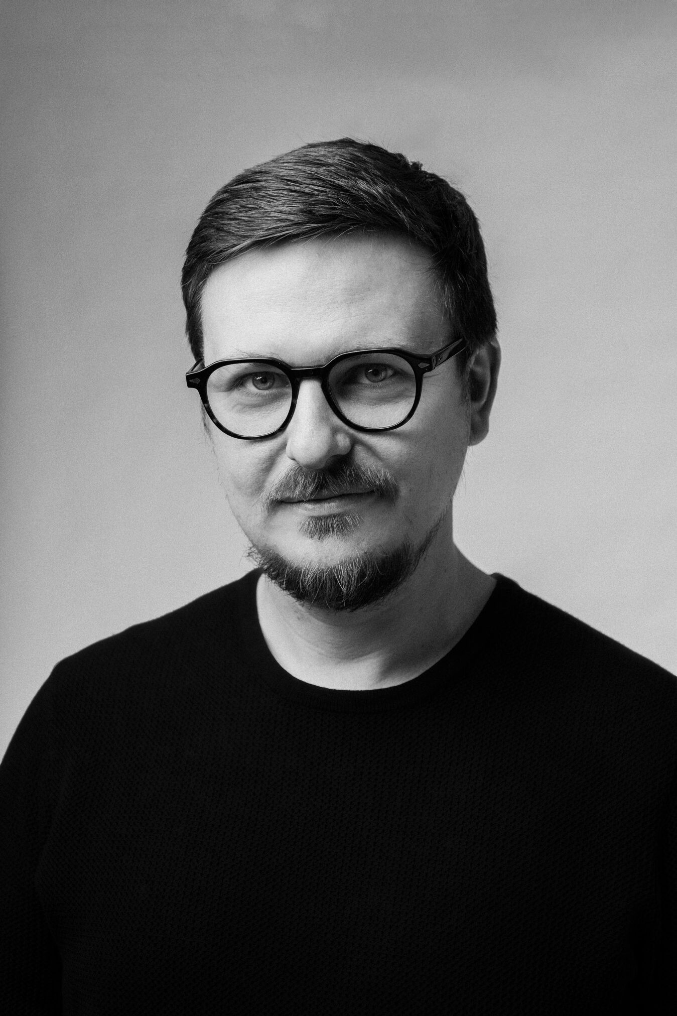Czarno-białe zdjęcie portretowe mężczyzny w okularach i czarnej koszulce, z wąsami i brodą kozią. Ma krótkie ciemne włosy i patrzy bezpośrednio w obiektyw na neutralnym tle.