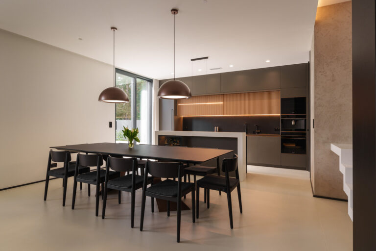Nowoczesna kuchnia połączona z jadalnią, wyposażona w czarny stół z sześcioma krzesłami oraz kuchnię z szarymi szafkami i drewnianą boazerią. Nad stołem wiszą dwie miedziane lampy wiszące, a całość utrzymana jest w minimalistycznym stylu z beżowo-szarą kolorystyką.