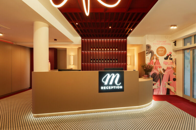 Nowoczesna recepcja hotelu z podświetlanym logo "m RECEPTION" na beżowym blacie, za którą znajduje się ściana z drewnianymi panelami i butelkami. Po prawej stronie widoczny jest kolorowy mural z postaciami kobiet w różowych strojach, a całe wnętrze utrzymane jest w ciepłej kolorystyce beżu i czerwieni.