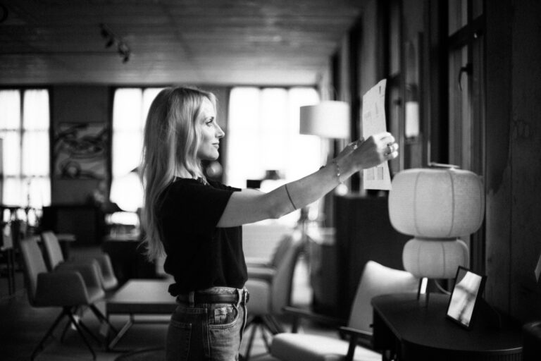 Czarno-biała fotografia przedstawiająca kobietę o długich blond włosach, która przygląda się lub dotyka elementu wyeksponowanego na drewnianej ścianie w przytulnym wnętrzu. W tle widoczne są duże okna wpuszczające naturalne światło, designerskie meble oraz lampy, tworzące ciepłą, domową atmosferę. Kadry pionowe i miękkie światło nadają scenie intymny, spokojny charakter. Autor zdjęcia: Radek Słowik bilbil fotografia.