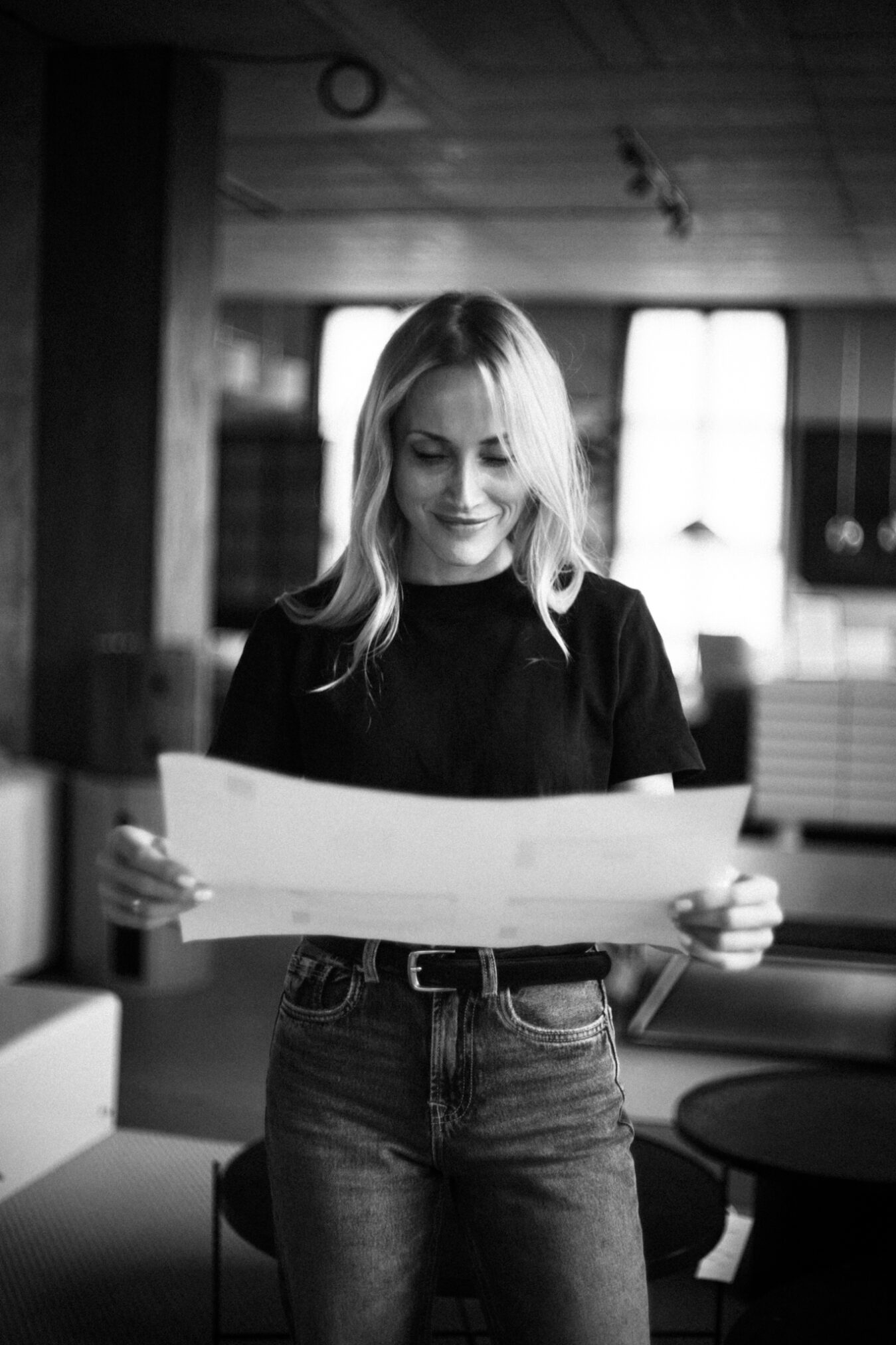 # Alt Text

Czarno-biała fotografia przedstawiająca uśmiechniętą kobietę o jasnych włosach, trzymającą w rękach duży arkusz papieru. Ubrana jest w czarną koszulkę i dżinsy z widocznym paskiem. W tle widać jasne, przestronne wnętrze z dużymi oknami wpuszczającymi światło, co tworzy spokojny, ciepły nastrój. Autor zdjęcia: Radek Słowik, bilbil fotografia.