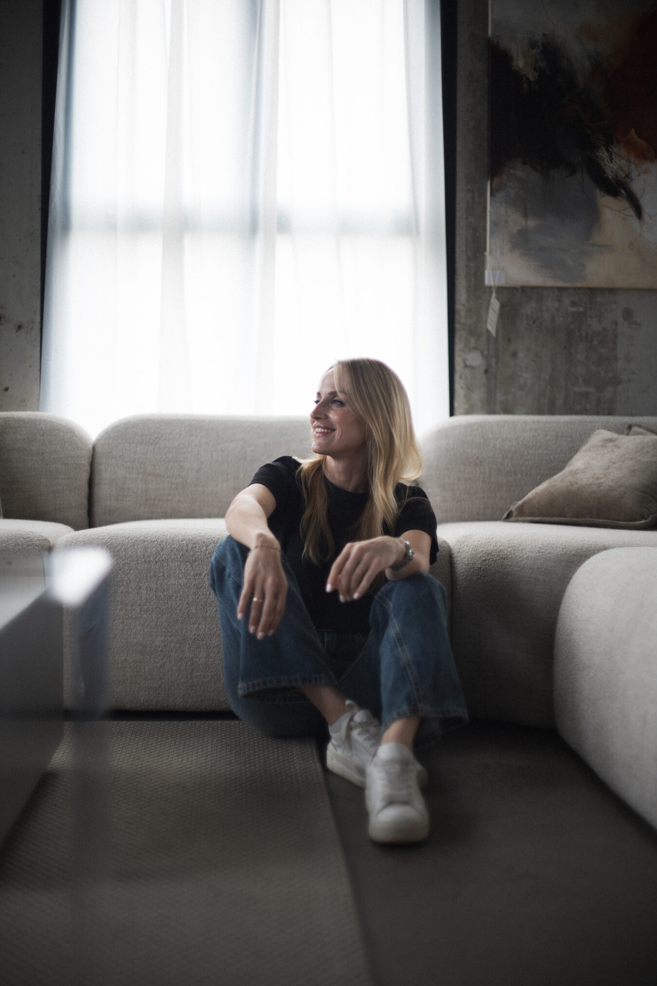 Kobieta o blond włosach siedzi zrelaksowana na jasnej, dwuosobowej sofie w industrialnym wnętrzu. Ubrana jest w czarny t-shirt, dżinsy i białe sneakersy, a jej uśmiech i swobodna pozycja sugerują poczucie komfortu i spokoju. Za nią widać duże okno wpuszczające miękkie, naturalne światło oraz surową betonową ścianę z abstrakcyjnym obrazem. Atmosfera zdjęcia jest ciepła i intymna, podkreślająca naturalność i luźność chwili. Autorem zdjęcia jest Radek Słowik bilbil fotografia.