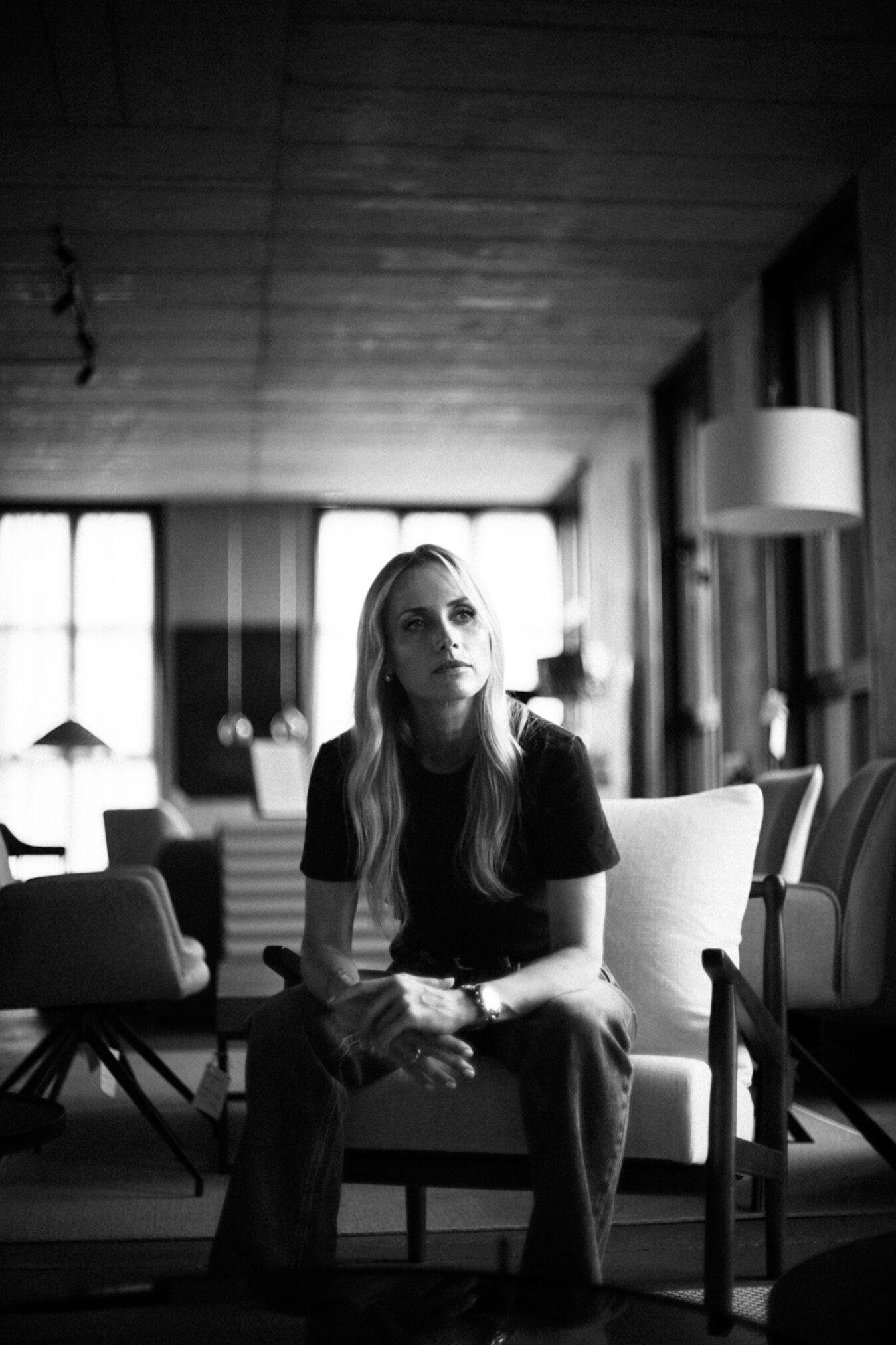 # Opis obrazu

Czarno-biała fotografia przedstawiająca młodą kobietę z długimi blond włosami, siedzącą na jasnej sofie w przestronnym salonie. Za nią widoczne są duże okna wpuszczające naturalne światło, drewniane belki na suficie oraz lampa podłogowa. Atmosfera zdjęcia jest kameralna i introspektywna, z wyraźnym kontrastem między ciemnymi elementami pierwszego planu a rozświetlonym tłem. Autor zdjęcia: Radek Słowik bilbil fotografia.