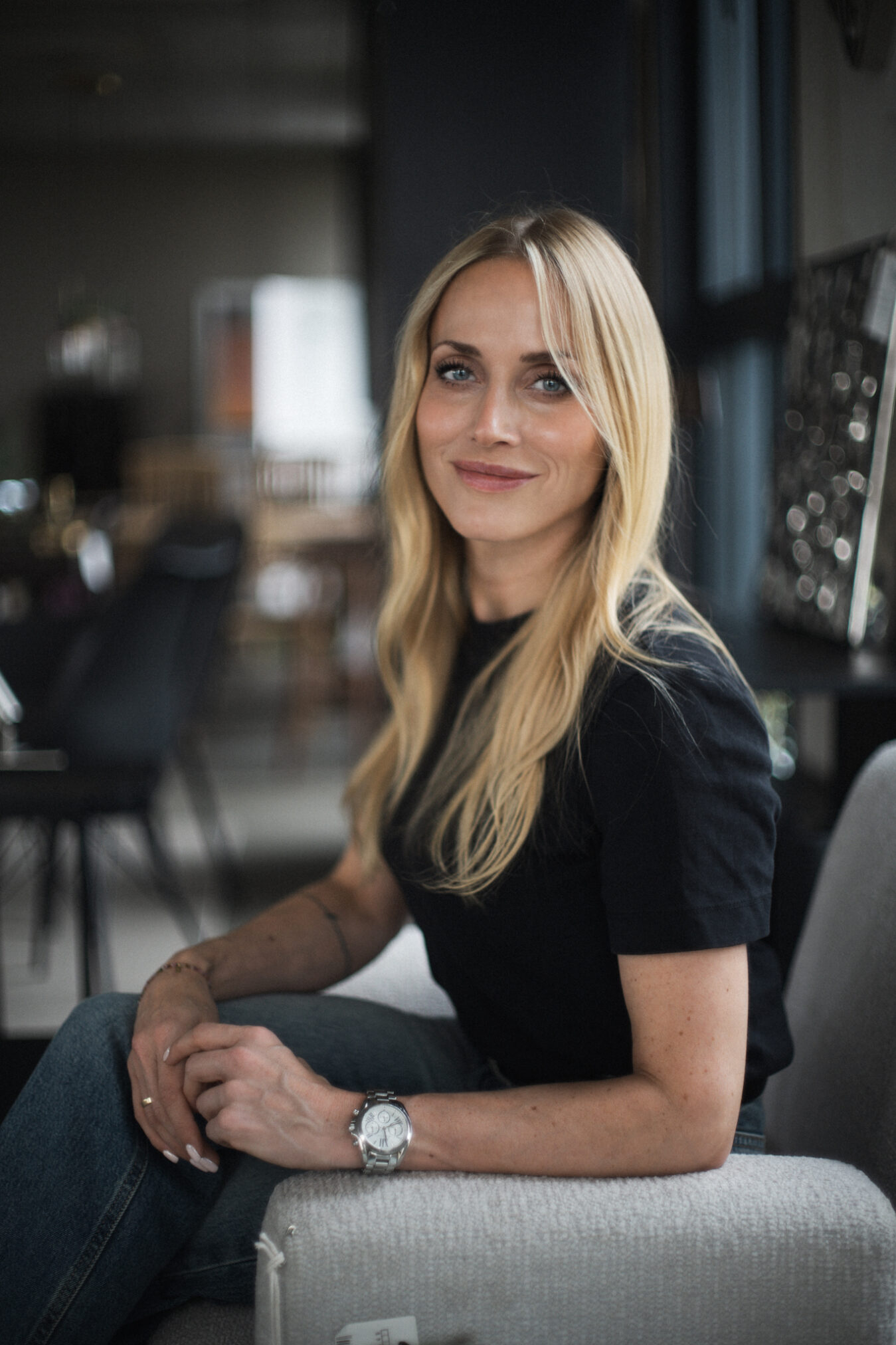 Kobieta o długich blond włosach siedzi na jasnej sofie w eleganckiej, ciemnej przestrzeni. Ma na sobie czarną koszulkę i zegarek na nadgarstku, patrzy spokojnie w obiektyw z subtelnym uśmiechem. W tle widać przytulne wnętrze z półkami i dodatkami, które tworzą kameralną, domową atmosferę. Autor zdjęcia: Radek Słowik bilbil fotografia.
