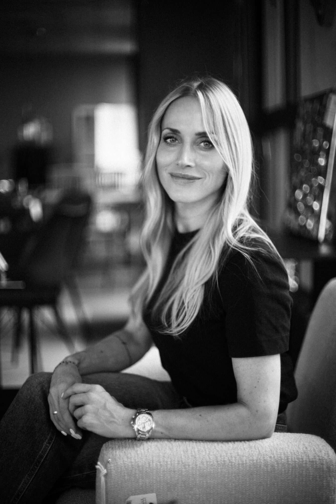 # Opis zdjęcia

Czarno-biała fotografia portretowa przedstawiająca uśmiechniętą kobietę o długich, jasnych włosach, siedząca przy stoliku w eleganckiej restauracji lub kawiarni. Kobieta ubrana w ciemną bluzkę, z delikatnym makijażem i zegarkiem na nadgarstku, patrzy bezpośrednio w obiektyw z ciepłym, przyjaznym wyrazem twarzy. W tle widać rozmyte wnętrze lokalu z miękkim, atmosferycznym oświetleniem i światełkami, co nadaje fotografii intymny, kameralny klimat. Autorem zdjęcia jest Radek Słowik bilbil fotografia.
