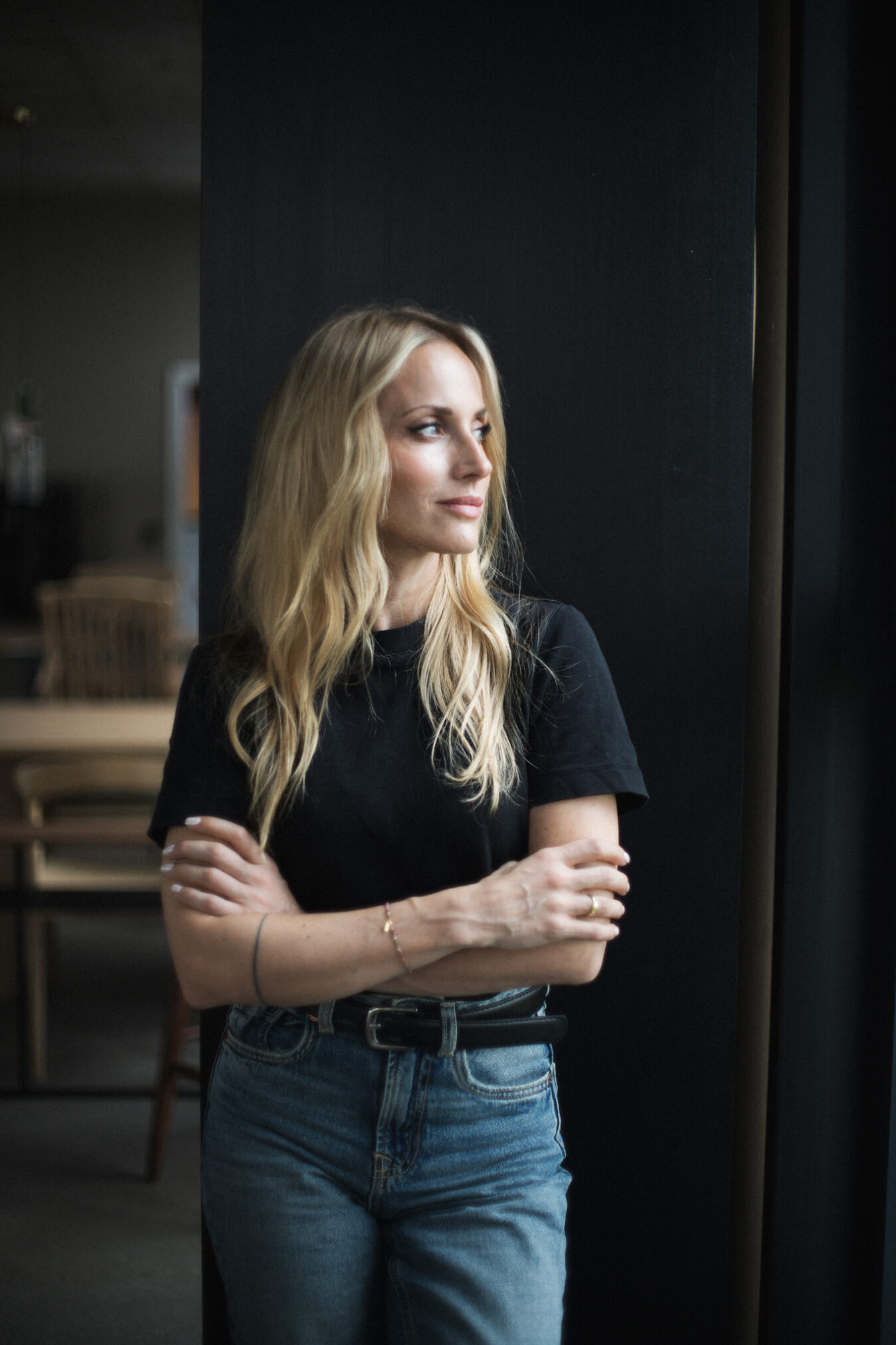 Kobieta z długimi blond włosami stoi przy ciemnej ścianie, patrząc w bok. Ma założone ręce i uśmiecha się delikatnie. Ubrana jest w czarny t-shirt i dżinsy z paskiem. W tle widać rozmazane wnętrze z drewnianymi meblami, a naturalne światło pada na jej twarz, tworząc kameralny, intimny nastrój. Autor zdjęcia: Radek Słowik bilbil fotografia.