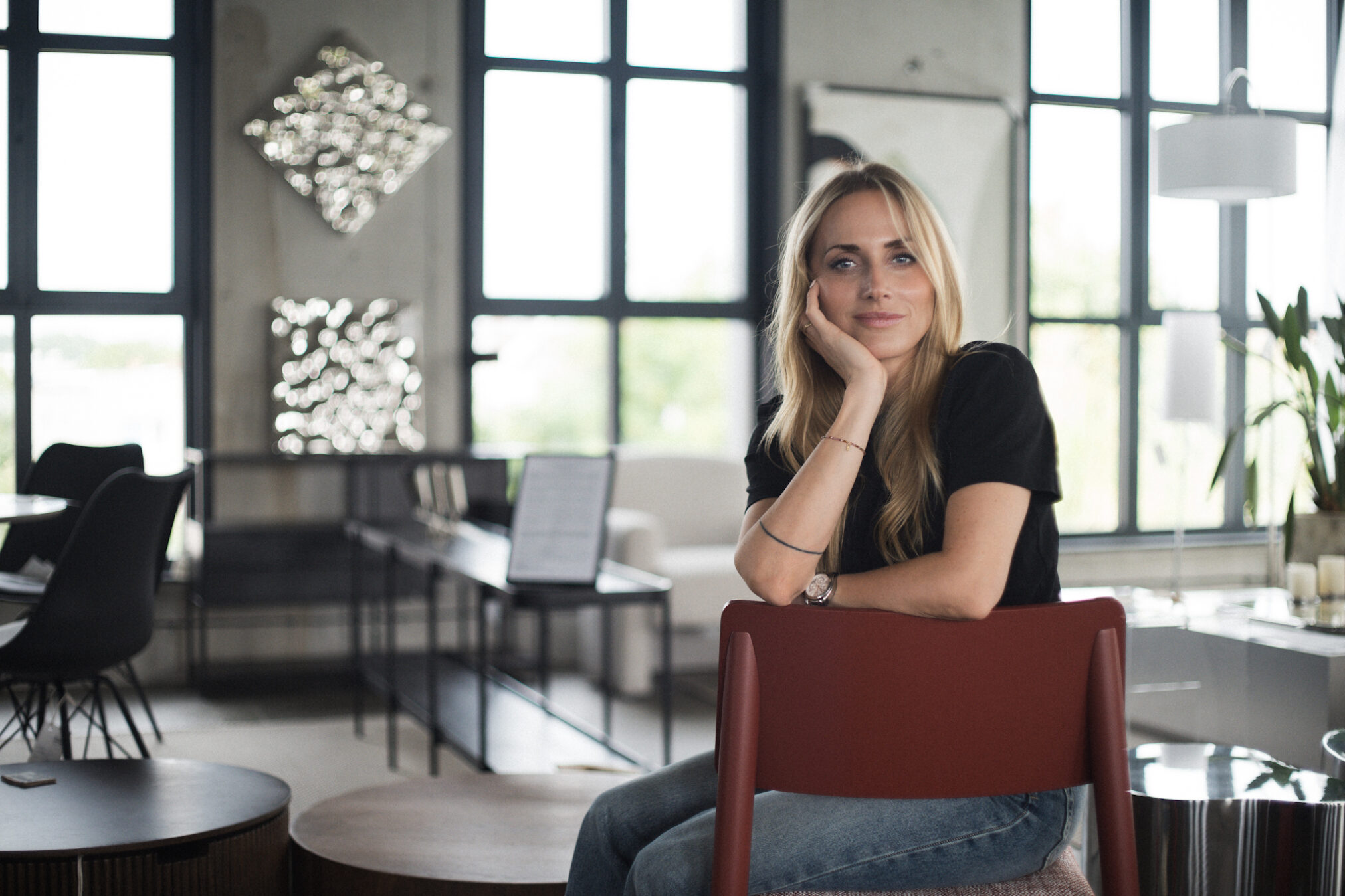 Kobieta o blond włosach siedzi na bordowym krześle przy stole w przestronnym wnętrzu w stylu industrialnym. Za nią widoczne są duże okna z czarnymi ramami wpuszczające naturalne światło oraz dekoracyjne elementy na ścianie. W pomieszczeniu panuje spokojna, profesjonalna atmosfera podkreślona szarymi betonowymi ścianami i minimalistycznymi meblami. Autor zdjęcia: Radek Słowik bilbil fotografia.